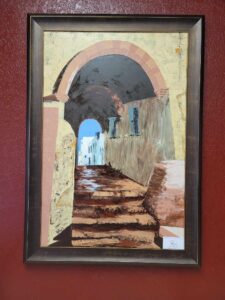 A framed painting of a Mediterranean architectural scene featuring a rustic stone staircase leading upwards through an arched tunnel, with light illuminating the passageway and buildings in the background against a reddish-brown wall.