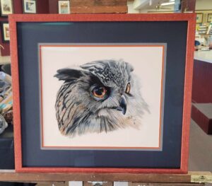 A framed painting of an owl with detailed features, including striking orange eyes and intricately painted feathers in shades of gray and brown. The painting has a dark blue mat with an orange border and is displayed on an easel in an art gallery.