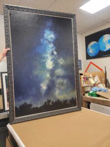 A person is holding a large framed painting depicting a night sky filled with stars and a nebula. The painting is set against a dark background with a silhouette of trees at the bottom. The scene appears to be in an art studio with various supplies and globes in the background.