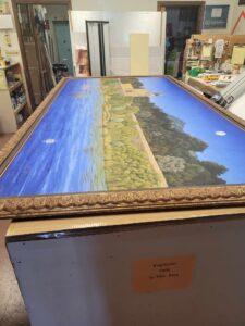A large, framed landscape painting sits on a table in a workshop. The painting depicts a serene scene with a lake, greenery, and a building in the distance under a blue sky. Tools and various workshop items are visible in the background. A sign reads "Employees Only In This Area.